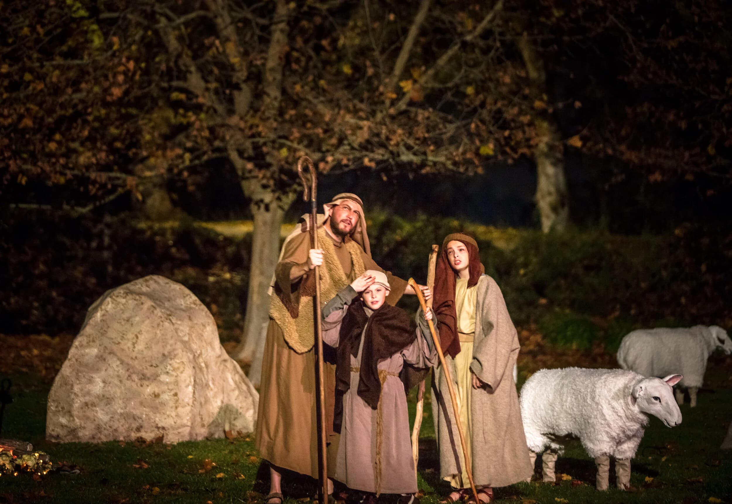 A shepherd and two children lean on wooden staffs beside sheep in a torchlit field at night.
