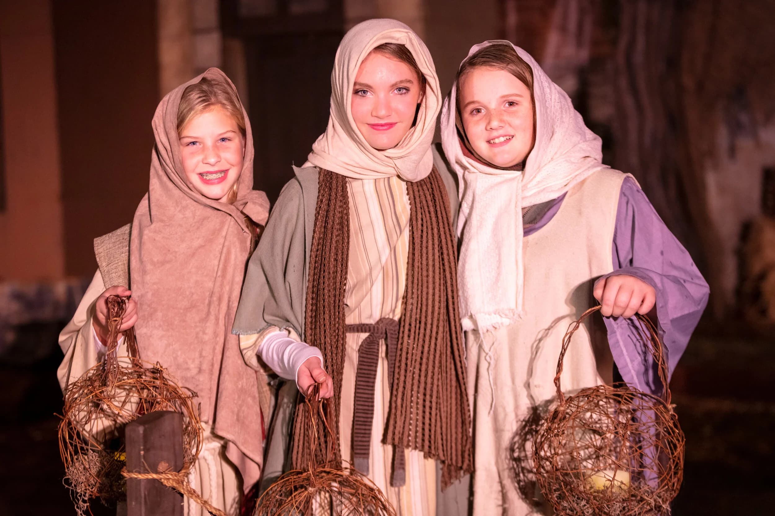 Three young women in headscarves hold woven basket lanterns and smile together at night.