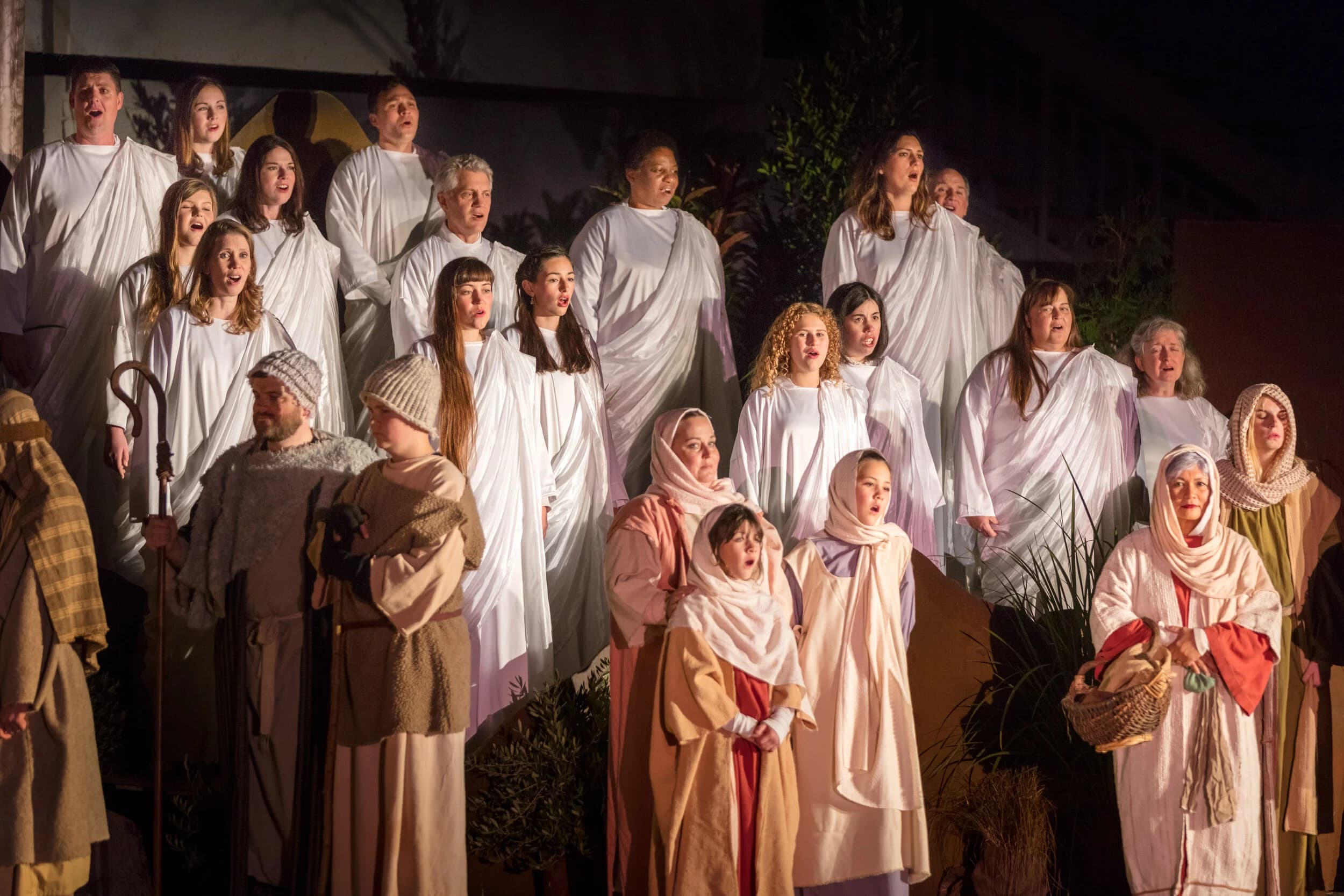 A large choir in white robes sings behind shepherds, women, and children gathered on an outdoor stage at night.